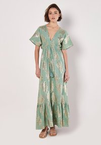 Robe longue vert menthe avec un décolleté en V profond, des manches courtes et des motifs de feuilles dorées. Dispose d'une taille à cordon et d'un ourlet en cascade.