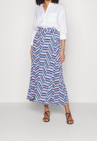 Maxihame valmistettuna kuvioidusta kankaasta, jossa on sinisiä, purppuran ja beigen värisiä sahalaita-motiiveja; yhdistetty valkoiseen napitettavaan paitaan ja mustiin sandaaleihin.