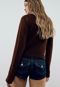 Jeune femme aux longs cheveux blonds, portant un pull marron et un short en denim bleu foncé avec des poches arrière boutonnées, se tenant de profil.