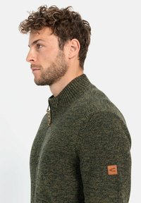 Grüner Strickpullover mit hohem, geripptem Kragen und halblanger Reißverschlusstaste, der einen Lederpatch-Akzent am linken Ärmel aufweist. Strukturiertes Material.