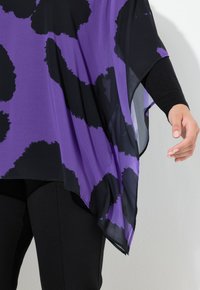 Violetti ja musta kuvioitu tunika, jossa on valuvia kankaita, epäsymmetrinen helma ja pitkät hihat, yhdistetty mustiin istuviin housuihin.