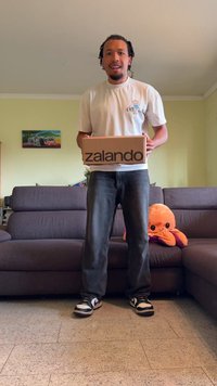 Une boîte en carton de la marque Zalando tenue par une personne portant un t-shirt blanc, un jean gris et des baskets noir et blanc, avec un pieuvre en peluche orange sur un canapé.