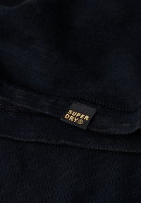 Tessuto blu navy con una texture sottile; presenta un'etichetta nera con lettering dorato "SUPER DRY" sul bordo, dettagli di orlo cuciti visibili.