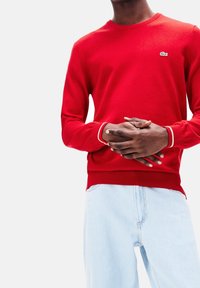 Persona che indossa una maglietta rossa a maniche lunghe con un piccolo logo di coccodrillo e jeans azzurri chiari, con le mani incrociate in vita su uno sfondo semplice.