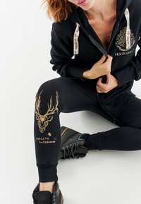 Čierna mikina na zips s emblémom Rokfortu, skombinovaná s čiernymi legínami s dizajnom zlatého Patronusa. Doplněné čiernymi členkovými topánkami.