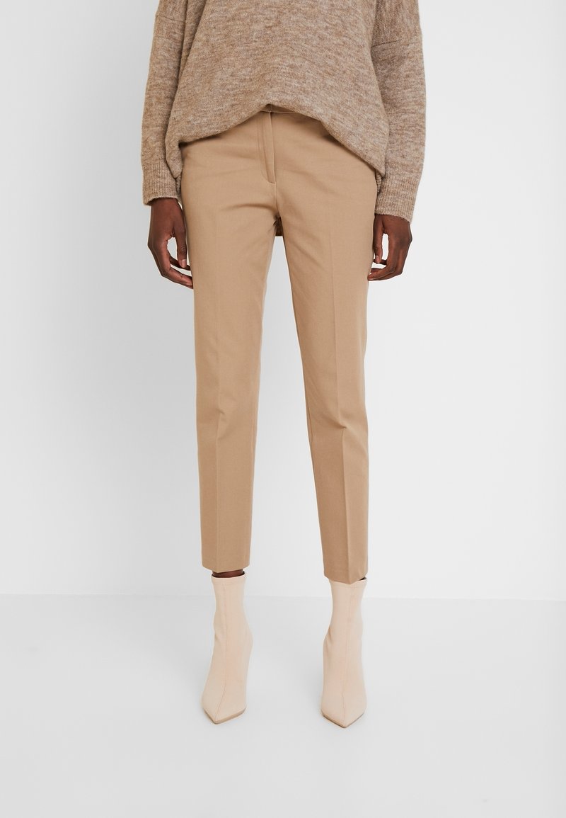 Personne portant un pantalon beige ajusté jusqu'à la cheville, des bottes beige clair à bout pointu et un pull en tricot marron ample sur fond blanc.