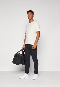Hellbeige T-Shirt mit gesticktem Logo, schwarze Sporthose und weiße Sneakers. Die Person hält eine schwarze Sporttasche. Glatte Texturen.