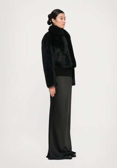 Woman wearing a black faux fur jacket and long black satin skirt, standing sideways against a plain white background.