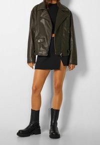 Olivgrön imitation läder bikerjacka med breda slag och blixtlås, i kombination med en svart minikjol och kraftiga svarta anklarstövlar.