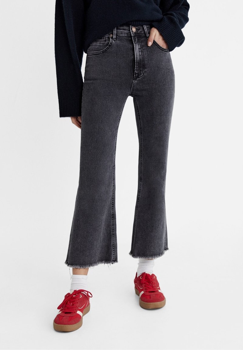 Personne portant un jean flare noir raccourci avec des ourlets bruts, des chaussettes blanches et des baskets rouges, debout avec une main dans la poche.