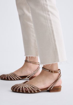 Pies con pantalones beige recortados y sandalias planas marrones con tiras con cuentas y hebillas en el tobillo sobre un fondo claro.