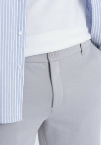 Helle graue Hose aus strukturiertem Stoff mit Knopfverschluss, kombiniert mit einem weißen Hemd unter einem blau gestreiften Button-Up.