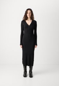 Black ribbed knit dress with long sleeves, V-neck, button detailing, and side slits; paired with black ankle boots featuring buckles.