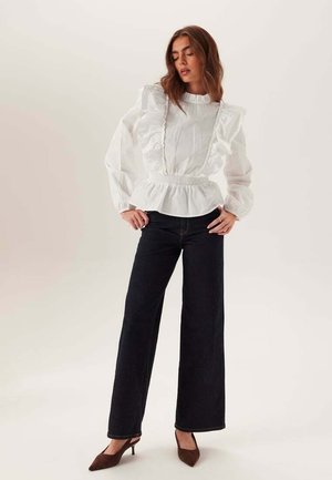 Vrouw met een witte blouse met lange mouwen en ruches, zwarte jeans met brede pijpen en bruine puntige hakken, staand met handen in de zij.