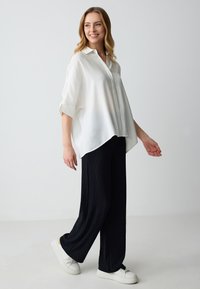 Camicia in lino bianco con una vestibilità rilassata, maniche corte e orlo curvo, abbinata a pantaloni larghi neri con texture e sneakers bianche.