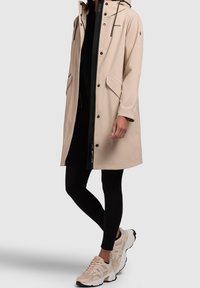 Imperméable beige avec capuche, fermeture éclair noire, boutons-pression et deux poches avant, assorti à un legging noir et des baskets légères.
