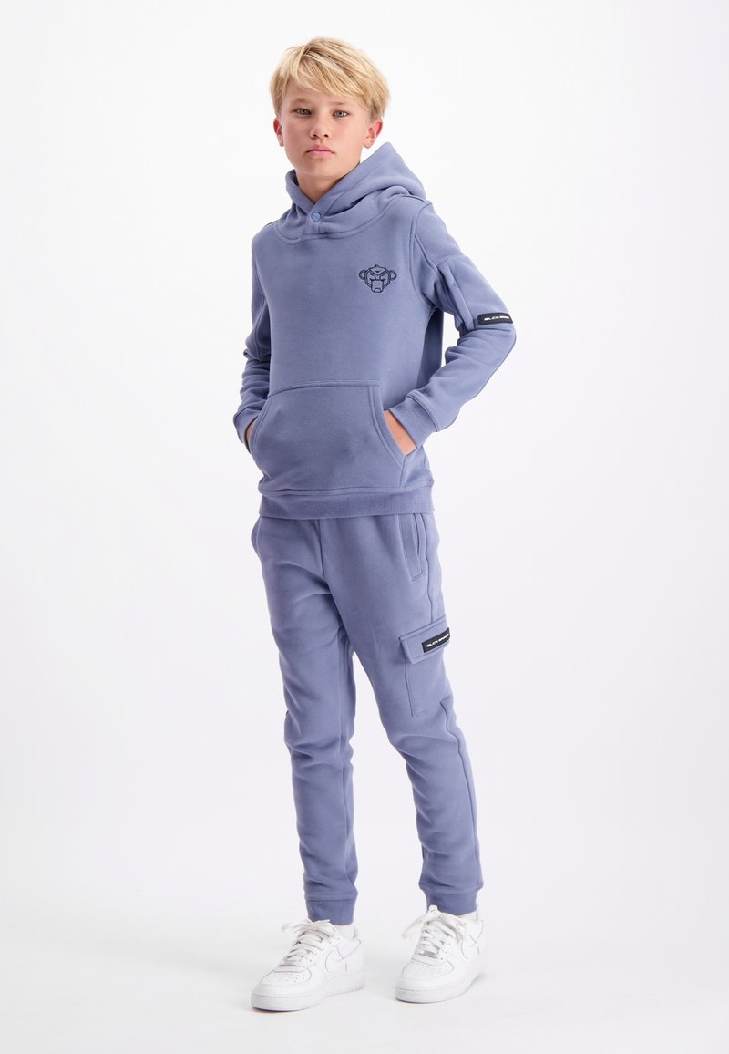 Blauw hoodie met kangoeroezak, logo op de borst en bijpassende tapered sweats met zijzak en zwarte accenten. Witte sneakers.
