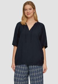 Navyblauwe blouse met een V-hals en een koord aan de onderkant, met korte mouwen en een ontspannen pasvorm. Gepaard met gemusterde, wijde broeken.