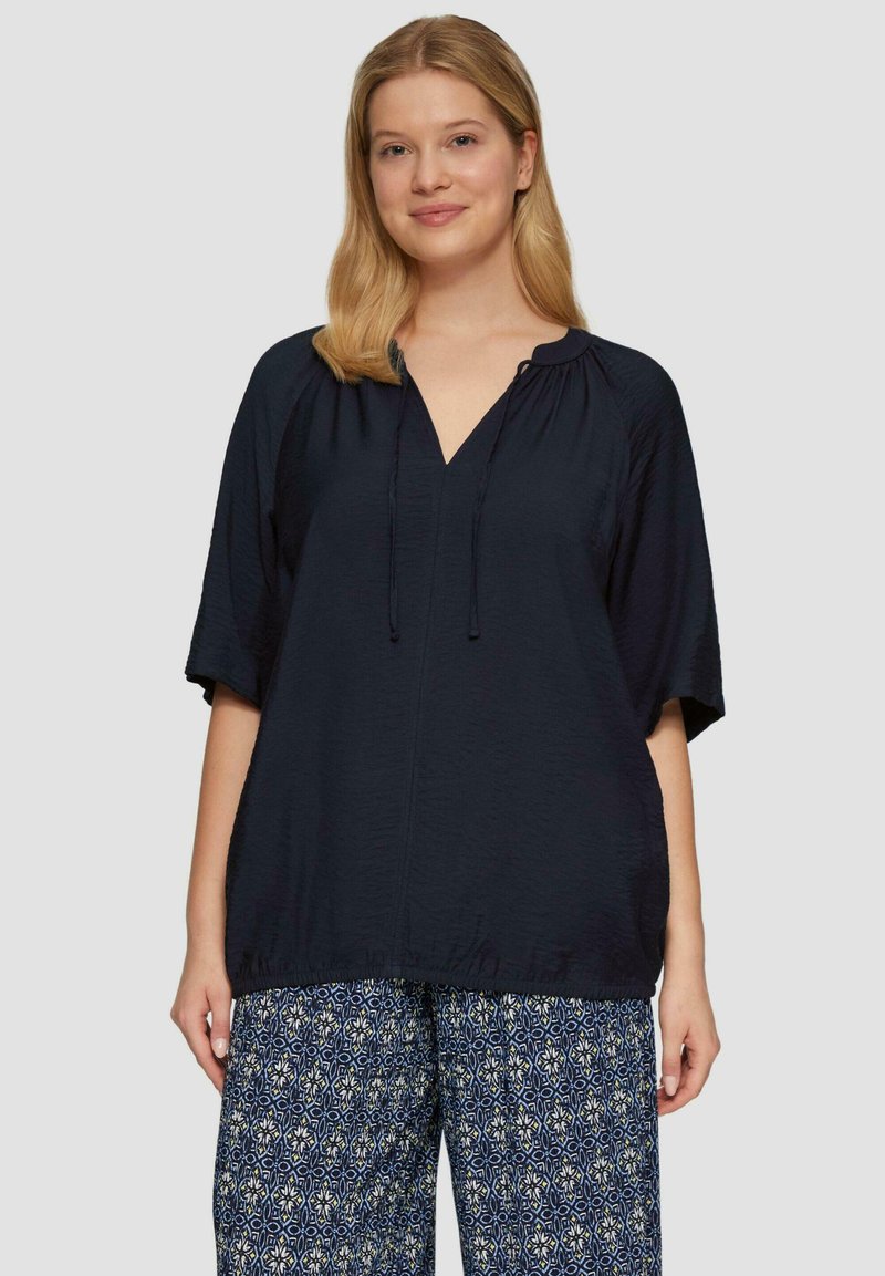 Navyblauwe blouse met een V-hals en een koord aan de onderkant, met korte mouwen en een ontspannen pasvorm. Gepaard met gemusterde, wijde broeken.
