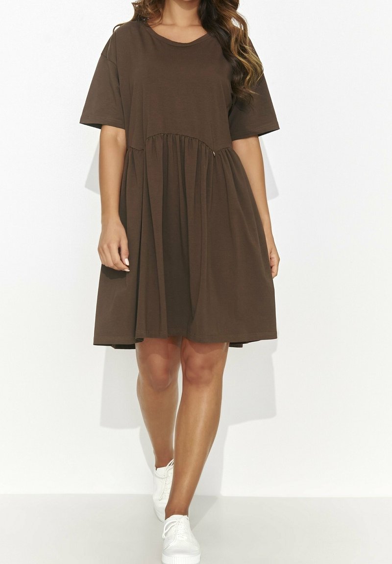 Femme portant une robe marron ample à manches courtes avec une taille froncée, associée à des baskets blanches, se tenant devant un fond blanc uni.