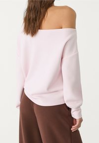 Rosa schulterfreier Pullover mit langen Ärmeln und lockerer Passform, gefertigt aus einem weichen, glatten Stoff, kombiniert mit braunen Hosen mit weitem Bein.