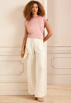 Donna con capelli ricci che indossa un top rosa senza maniche e pantaloni bianchi a gamba larga, che tiene una borsa beige, in piedi all'interno davanti a una parete a pannelli.