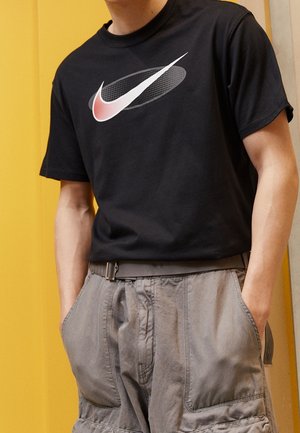 Czarna, krótka koszulka z krótkim rękawem z dużym logo Nike w kolorze czerwonym i białym. Połączona z szarymi szortami cargo. Gładki materiał bawełniany.