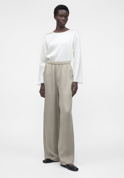 Γυναίκα που φοράει μπεζ παντελόνια με φαρδιά μπατζάκια, λευκό πουκάμισο με μακριά μανίκια και μαύρα επίπεδα παπούτσια, στέκεται μπροστά σε απλό φωτεινό φόντο.