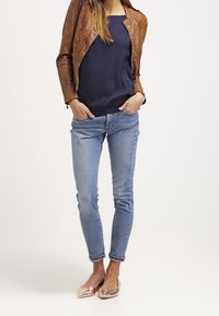 Giacca di pelle marrone sopra una maglietta blu navy, abbinata a jeans skinny azzurri e ballerine nude lucide. Sfondo semplice.