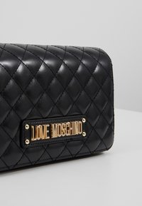 Pehmustettu musta keinonahkainen lompakko, jossa on kultaisen värinen laatta, jossa lukee "LOVE MOSCHINO." Lompakossa on vinot tikkauskuviot ja minimalistinen muotoilu.