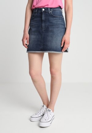 Mini falda de mezclilla en azul oscuro con un corte ajustado, que cuenta con un cierre de botón y cremallera, y un dobladillo sin acabar. Combinada con zapatillas blancas.