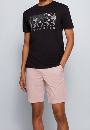 Mand iført sort Hugo Boss T-shirt med geometrisk print og lyserøde shorts til knæet, stående med den ene hånd i lommen.
