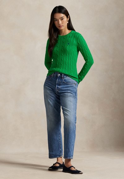 Femeie cu păr lung și închis la culoare, purtând un pulover verde cu model împletit, blugi albaștri cu talie înaltă și pantofi negri Mary Jane, stând cu mâinile pe spate.