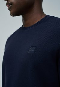 Hombre vistiendo una camisa de manga larga texturizada de color azul oscuro con un parche de "Salsa Jeans" en el pecho izquierdo, mostrado desde el cuello hasta la parte superior del torso.