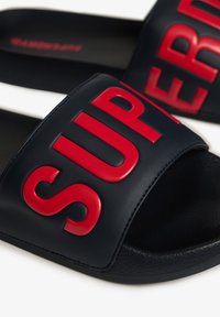 Sandales à glissière bleu marine avec une semelle noire texturée. Lettres rouges en gras "SUPER" sur la bride. Fabriquées en matériau synthétique lisse.