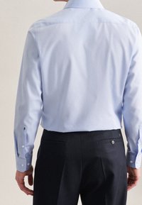 Camicia azzurro chiaro con maniche lunghe, polsi a bottoni e colletto tradizionale, abbinata a pantaloni scuri su misura. Texture liscia, dettagli minimi.