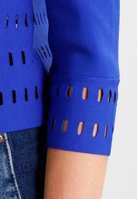 Blå stickad tyg med jämnt fördelade rektangulära utskärningar på ärmarnas manschetter och sidan, bärs med blå denimjeans.