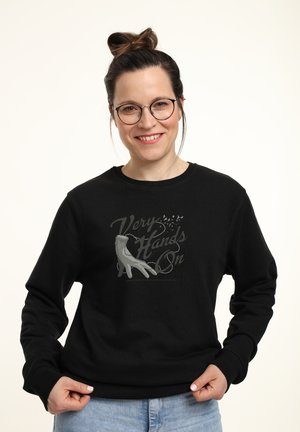 Donna sorridente con occhiali e chignon che indossa una felpa nera con una grafica di una mano e il testo 'Very Hands On', mentre tiene l'orlo, su uno sfondo chiaro.