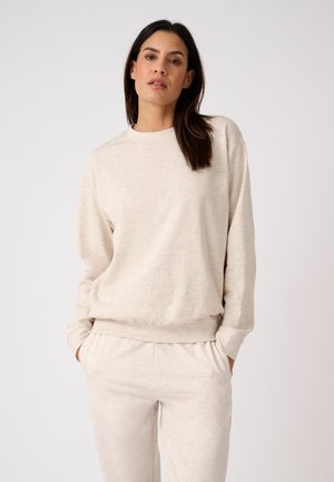 Donna con capelli scuri che indossa una felpa beige chiaro e pantaloni abbinati con le mani nelle tasche su uno sfondo chiaro e uniforme.