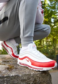 Le scarpe Nike Air Max 97 bianche e rosse presentano un design ondulato, materiali in pelle e rete, un'imbottitura ad aria visibile e un piccolo logo decorativo.