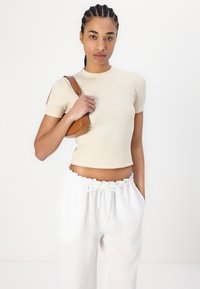 Top corto a maniche corte ribbed di colore crema chiaro con design aderente, abbinato a pantaloni bianchi con cordoncino e una piccola borsa marrone.