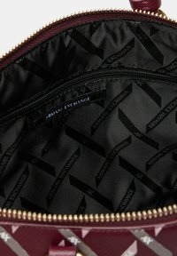 Interno della borsa a tracolla color bordeaux con una tasca con zip, foderato in tessuto nero caratterizzato da un motivo ripetuto del logo Armani Exchange. Dettaglio della zip dorata visibile.
