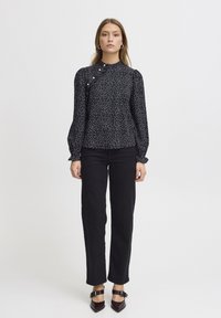 Blusa nera con pois bianchi, colletto alto e maniche lunghe a sbuffo. Abbinata a pantaloni neri a gamba dritta e scarpe scure a punta.