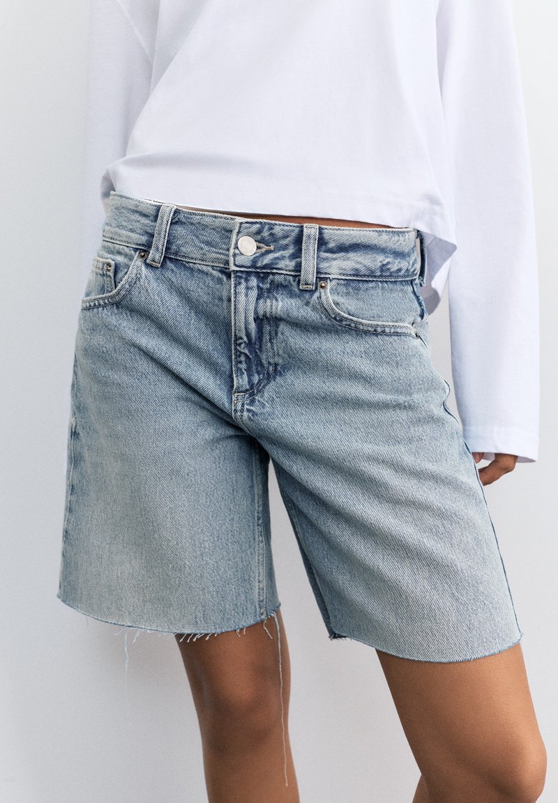 Person wearing light blue denim shorts with frayed hems and a white long-sleeve shirt, shown from mid-thigh to waist.