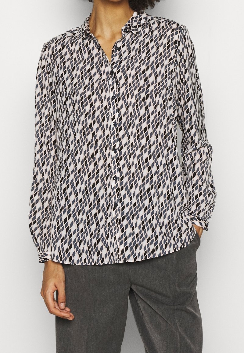 Femme portant un chemisier boutonné à manches longues avec un motif losanges noir et blanc et un pantalon gris, une main dans la poche.