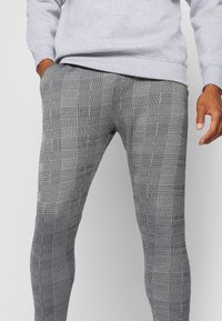 Osoba w szarych i czarnych spodniacach w kratkę slim-fit z rękami w pobliżu kieszeni, uzupełniona jasnoszarym sweatshirt'em.