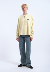 Gelbes Langarmhemd mit "good vibes" Grafik, kombiniert mit hellblauen Weitbeinejeans und schwarzen Sneakers. Vor einem schlichten Hintergrund.