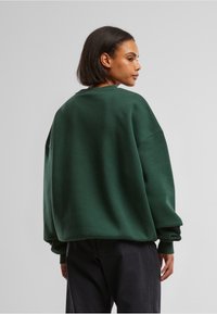 Felpa oversize con girocollo in verde scuro, realizzata in tessuto morbido con colletto e polsini a costine, caratterizzata da spalle basse e una vestibilità rilassata.