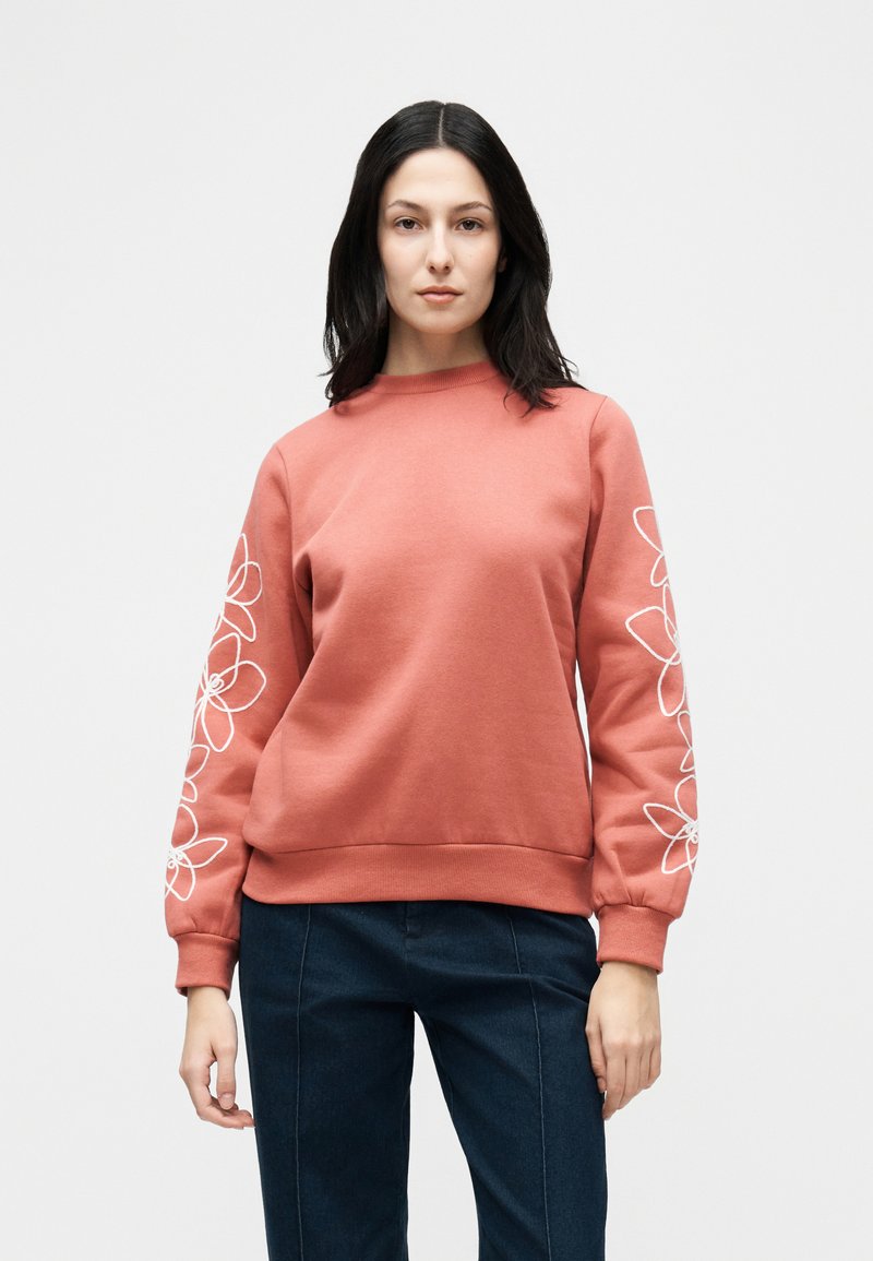 Γυναίκα με σκούρα μαλλιά που φοράει κοραλλί φούτερ με λευκή κεντημένη φλοραλ λεπτομέρεια στα μανίκια και σκούρο μπλε παντελόνι, στέκεται μπροστά σε απλό φόντο.