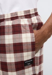 Rote und cremefarbene karierte Shorts mit elastischem Bund, Seitentasche und schwarzem Etikett mit weißem Text in der Nähe des Saums. Glatte Stoffstruktur.